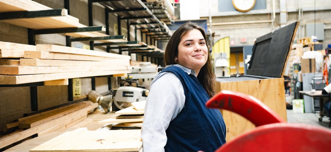 Avery McGarry stands in the scene shop among shelves and looks seriously at the camera.